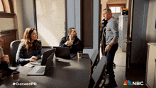 a group of people sitting around a table with laptops and a nbc logo on the bottom