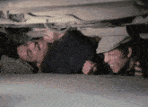 two men are laying under a car and one is wearing a flat cap