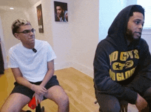 a man wearing a no cys hoodie sits next to a man wearing shorts