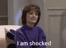 a woman in a purple sweater is sitting on a couch and saying i am shocked