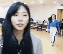 a woman with long black hair is standing in a room looking at the camera while another woman stands behind her .