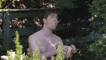 a shirtless man is peeling a banana in a garden while sitting on a bench .