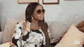 a woman wearing sunglasses is sitting on a couch next to a pillow .