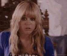 a woman with blonde hair and bangs is sitting in a chair making a surprised face .