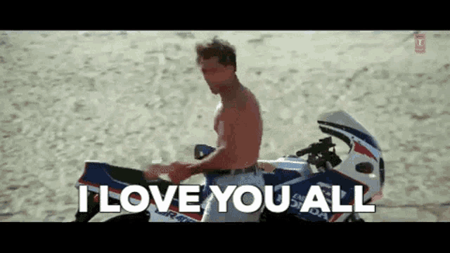 a man without a shirt is sitting on a motorcycle on the beach and saying i love you all .