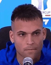 a man in a blue shirt is sitting in front of a microphone with a supercup logo behind him