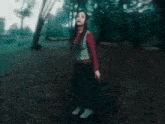 a woman in a red sweater and black skirt is standing in a forest .