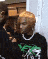 a man with blonde hair and a necklace is standing next to another man in a black shirt .