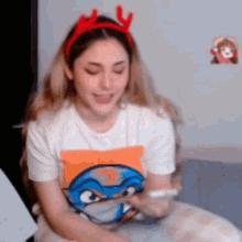a woman wearing a turtle t-shirt and reindeer antlers is sitting on a bed looking at her phone .