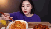 a woman in a purple shirt is eating a bowl of food with chopsticks