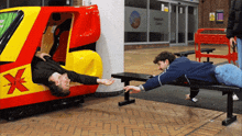 a man is laying on a bench next to a yellow and red vehicle with a red x on it