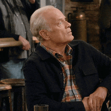 an older man wearing a plaid shirt and a black jacket sits at a table
