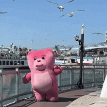 a pink teddy bear is standing on a pier with seagulls flying around him