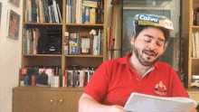a man wearing a red shirt is reading a book and has a calvo box on his head