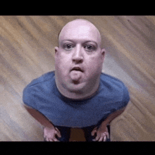 a bald man in a blue shirt is sticking his tongue out while standing on a wooden floor .