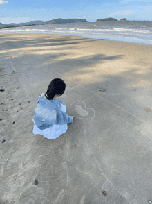 a girl is sitting on the beach drawing a heart in the sand