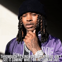 a man with dreadlocks wearing a black beanie and a purple jacket is holding his hand to his chin .