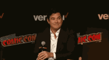 a man in a suit is sitting in front of a comic con sign