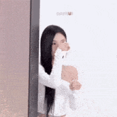 a woman with long black hair is covering her face with her hand while standing next to a wall .