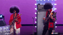two men with afros playing guitars and singing into microphones on a stage with a nbc logo in the background