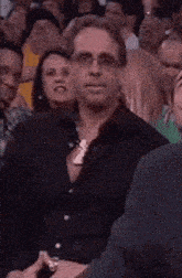 a man wearing glasses and a ring is sitting in a crowd of people and making a funny face .
