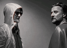 two men standing next to each other with one wearing a hood