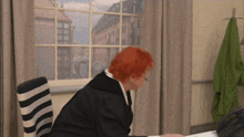 a woman with red hair sits at a desk in front of a window looking out