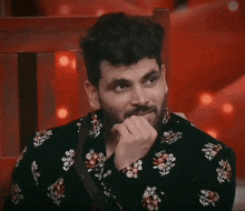 a man with a beard is wearing a black shirt with flowers on it and has his hand on his chin .