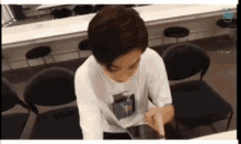 a young man in a white shirt is sitting at a table looking at a phone .