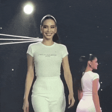 a woman wearing a white miss universe t-shirt walks down a runway