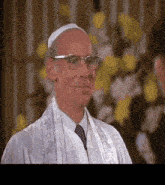 a man wearing glasses and a white robe stands in front of flowers