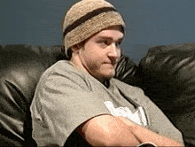 a man wearing a beanie and a grey shirt with the letter m on it sits on a couch
