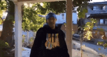 a man wearing a black hoodie and a blue hat is standing on a porch looking out a window .