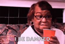 a woman with glasses is sitting on a couch holding a red cup and saying `` i be damned '' .