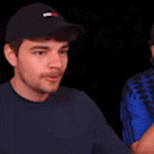 two men are standing next to each other and one of them is wearing a hat and a blue shirt .