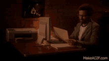 a man is sitting at a desk with a computer and a printer and a makeagif.com watermark