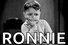 a little boy is sitting at a table with his hand on his chin and the word ronnie written on the screen .
