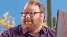 a man with a beard wearing glasses is sitting in front of a computer monitor .