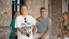 a woman wearing a shirt that says oh yes stands in front of a group of people