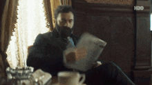 a man with a beard is reading a newspaper while sitting at a table .