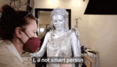a woman wearing a mask is standing next to a statue that says " i 'm a not smart person "