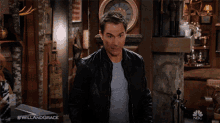 a man wearing a black leather jacket is standing in front of a fireplace and a clock with the hashtag #willandgrace