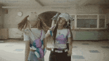 two girls are standing next to each other in a room . one of the girls is wearing a backpack that says oxygen .
