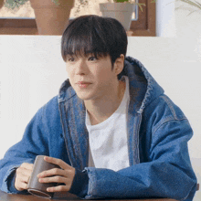 a young man in a denim jacket is sitting at a table holding a mug