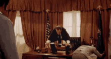 a man in a suit is kneeling down in front of a desk while another man stands behind him