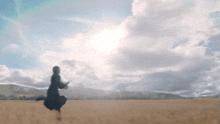 a woman in a black dress is running through a field with the sun shining through the clouds