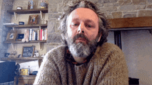 a man with a beard wearing a sweater is sitting in front of a fireplace
