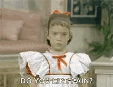 a little girl in a white dress is standing in a living room and saying `` do you like pain '' .