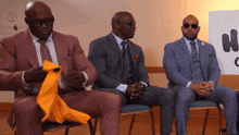 three men in suits are sitting in front of a sign that has the letter h on it