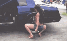 a woman is squatting down in front of a blue car holding a tire .